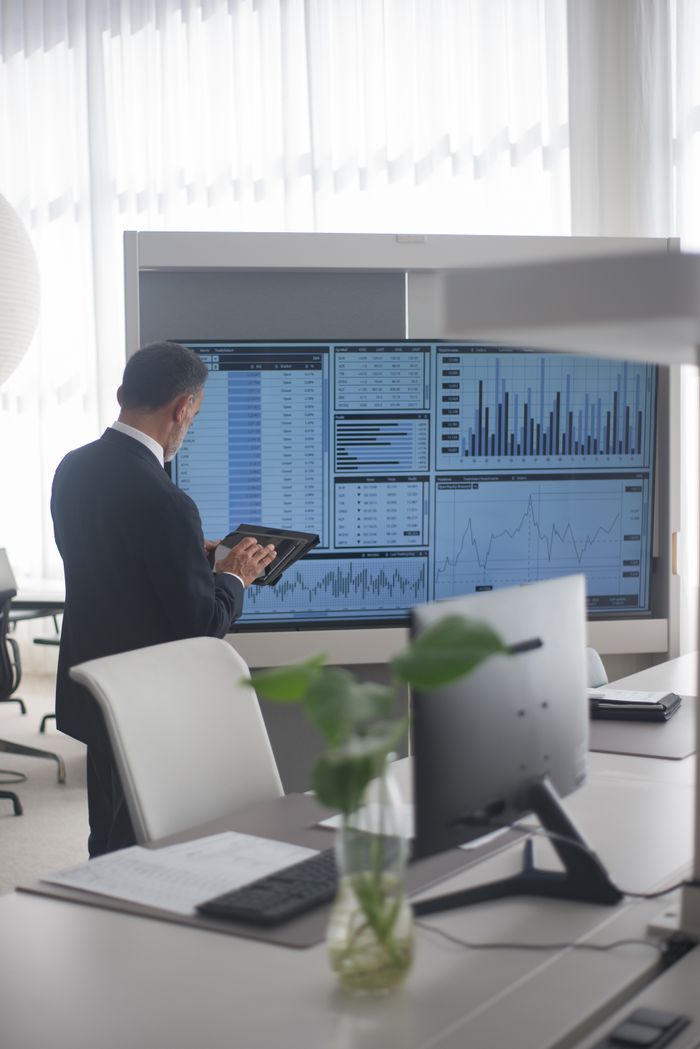 who-we-are A businessman in a modern office reviews data charts on a large screen with a tablet.